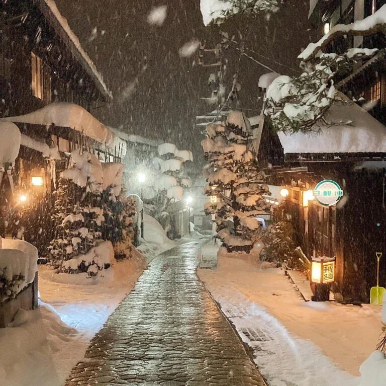 Snowy, lantern-lit street at night with a cobblestone path and snow-covered trees and buildings on both sides.