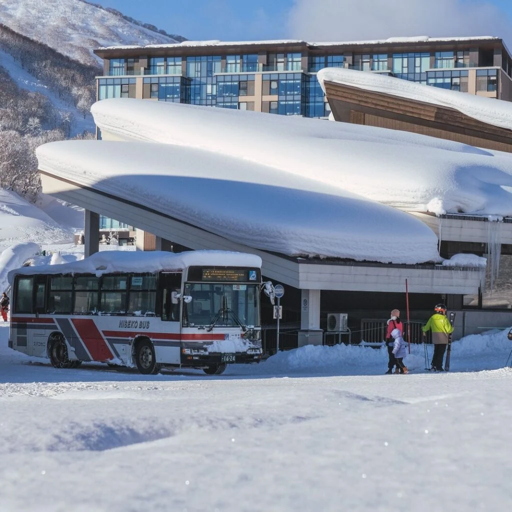 Snowy ski resort with a bus parked beside a modern building; people with skis and poles nearby, ready to depart or arrive.